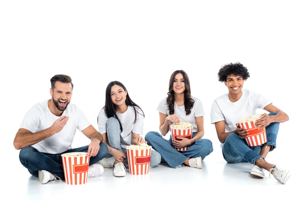 joyful multicultural friends sitting with popcorn buckets and watching movie on white - Фото, изображение