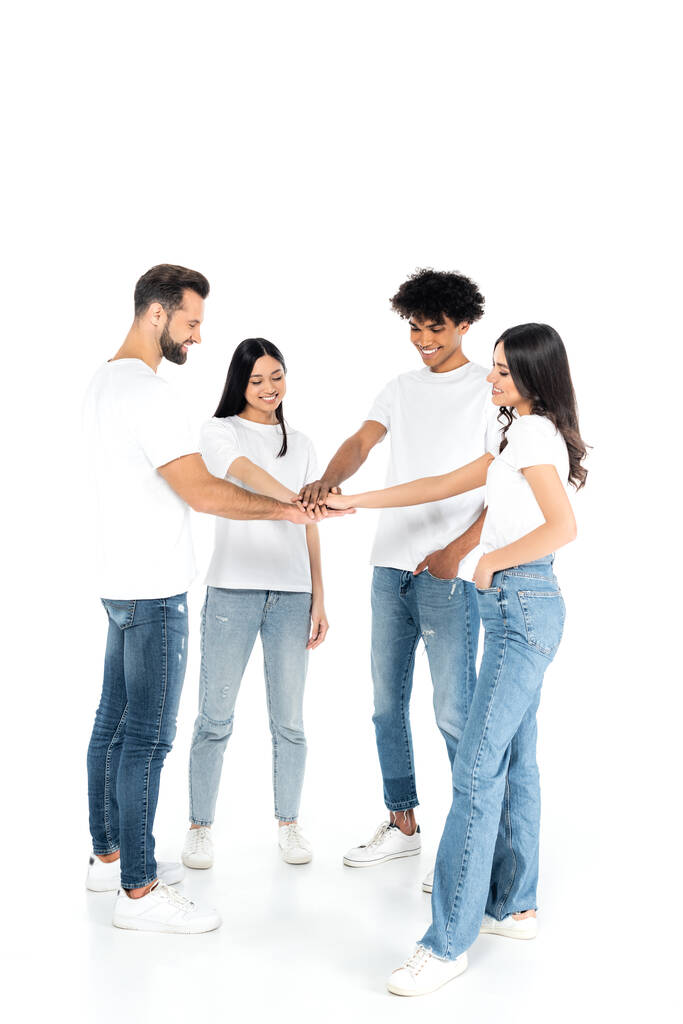 volledige weergave van multiculturele vrienden in t-shirts en jeans die de handen ineenslaan terwijl ze op wit staan - Foto, afbeelding