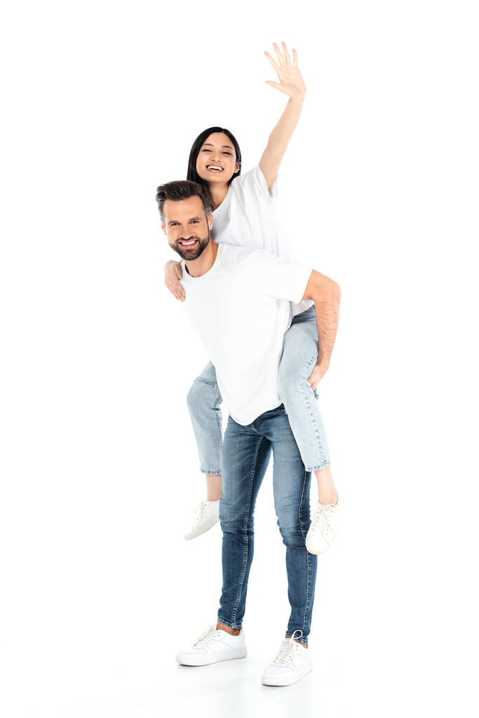 man in jeans piggybacking excited asian woman waving hand on white - 写真・画像