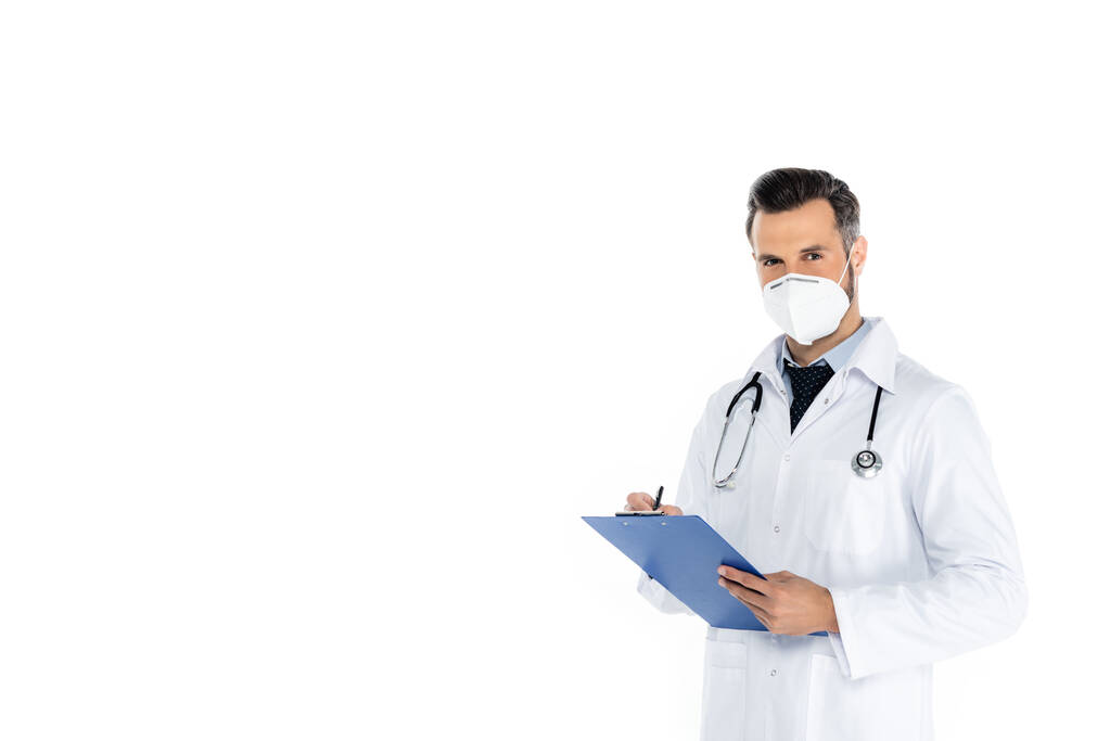 γιατρός με λευκό παλτό και ιατρική μάσκα γραφής στο πρόχειρο και κοιτάζοντας κάμερα που απομονώνονται σε λευκό - Φωτογραφία, εικόνα