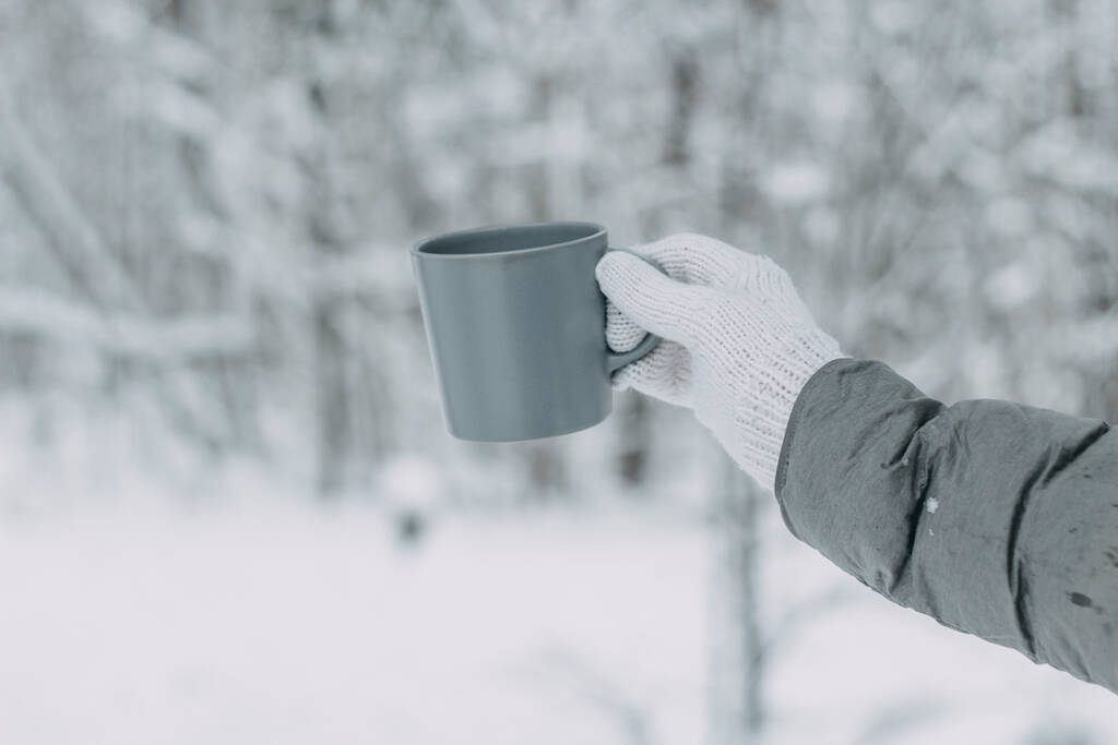 Een hand in een witte gebreide wollen handschoen en een grijze jas houdt een grijze mok tegen de achtergrond van een prachtig besneeuwd bos. - Foto, afbeelding