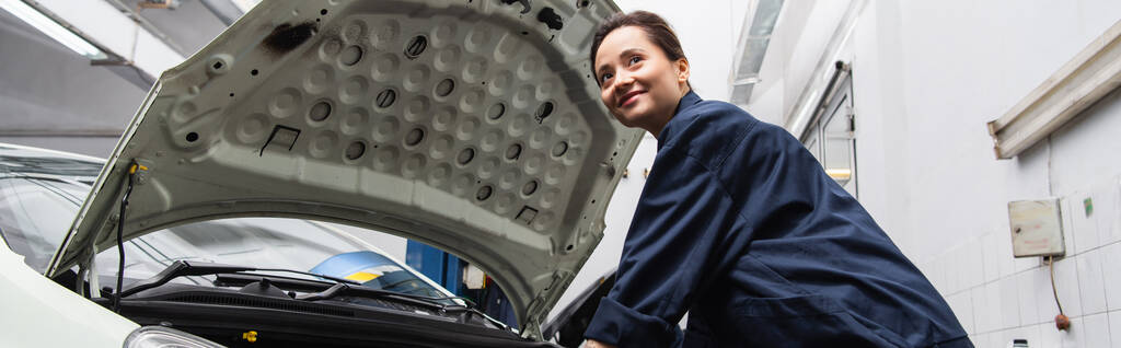 Positive mechanic looking away near car with open hood in garage, banner  - Foto, Imagem