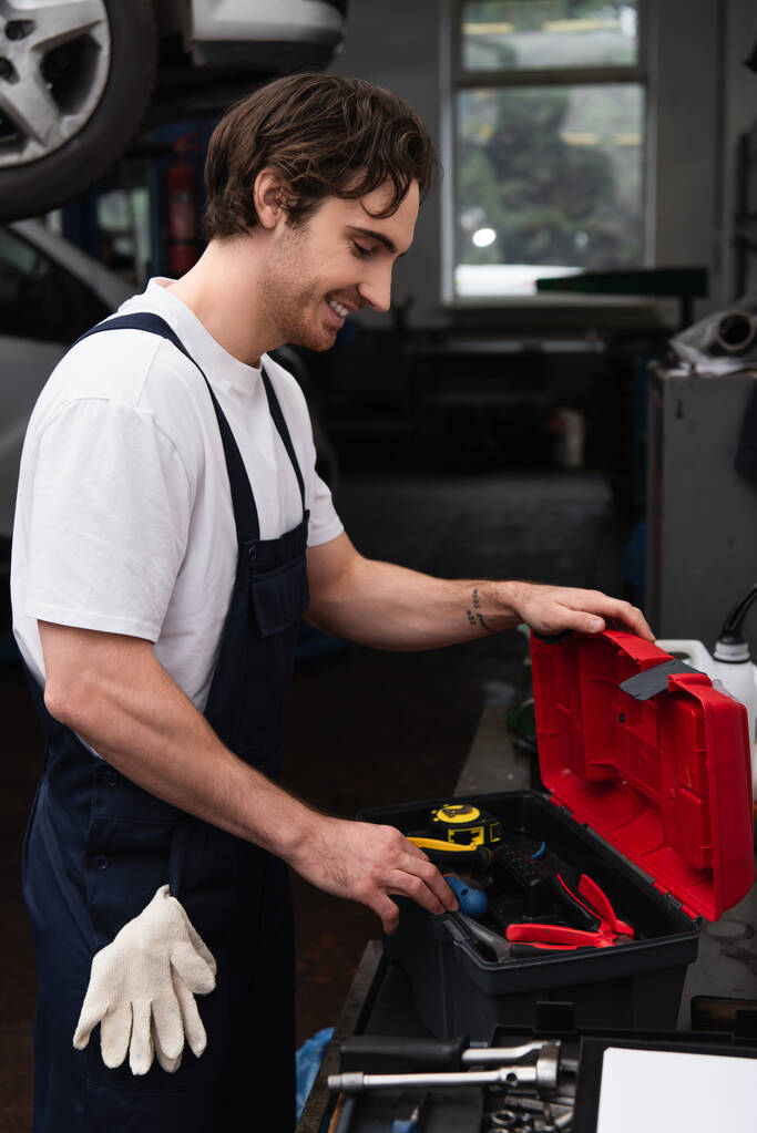 Side view of smiling workman looking at tools in toolbox in car service  - Фото, изображение