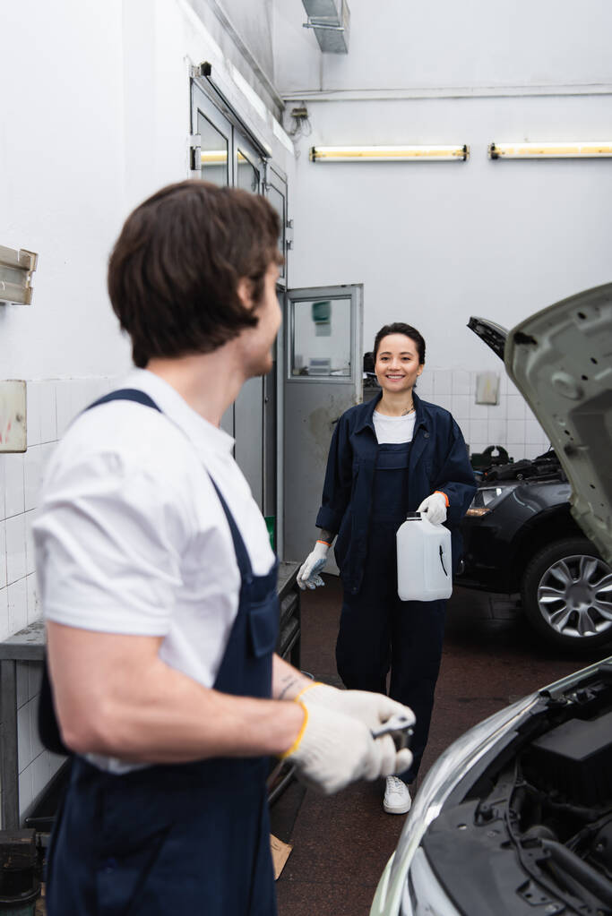 Smiling mechanic holding canister near blurred colleague and car in service  - Foto, Imagem
