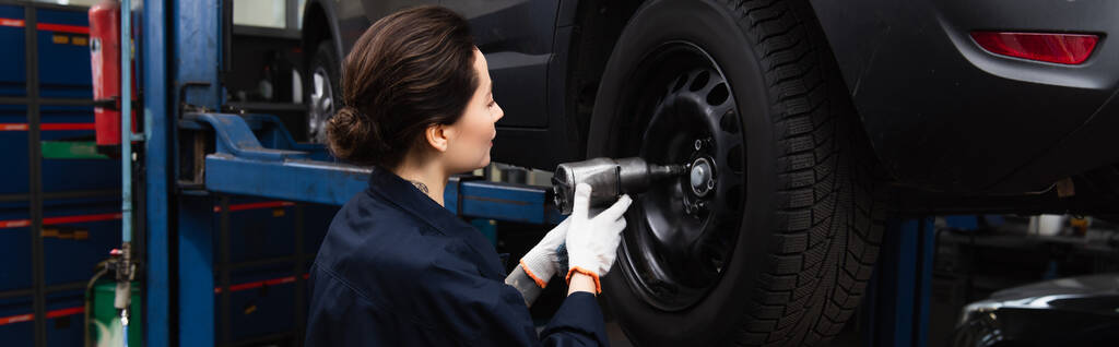 Side view of forewoman working with impact wrench and car wheel in garage, banner  - 写真・画像
