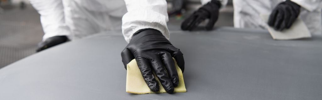 Przycięty widok robotnika w kombinezonie ochronnym i rękawiczkach przy użyciu papieru ściernego na masce samochodowej w garażu, baner  - Zdjęcie, obraz