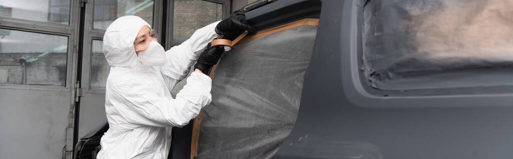 Trabajadora en traje protector y guantes aplicando cinta adhesiva en el coche en el garaje, pancarta  - Foto, Imagen