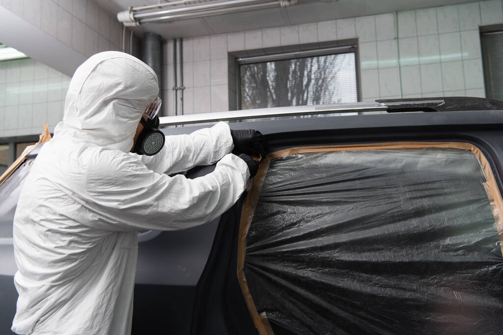 Werkman in masker en hazmat pak aanbrengen tape op cellofaan op auto in dienst  - Foto, afbeelding