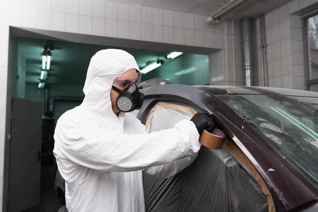 Workwoman in respirator and goggles applying tape on cellophane while working in car service  - Foto, Imagem