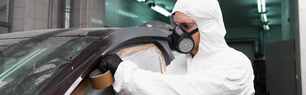 Young workman in hazmat suit applying tape on car with cellophane in garage, banner  - Zdjęcie, obraz