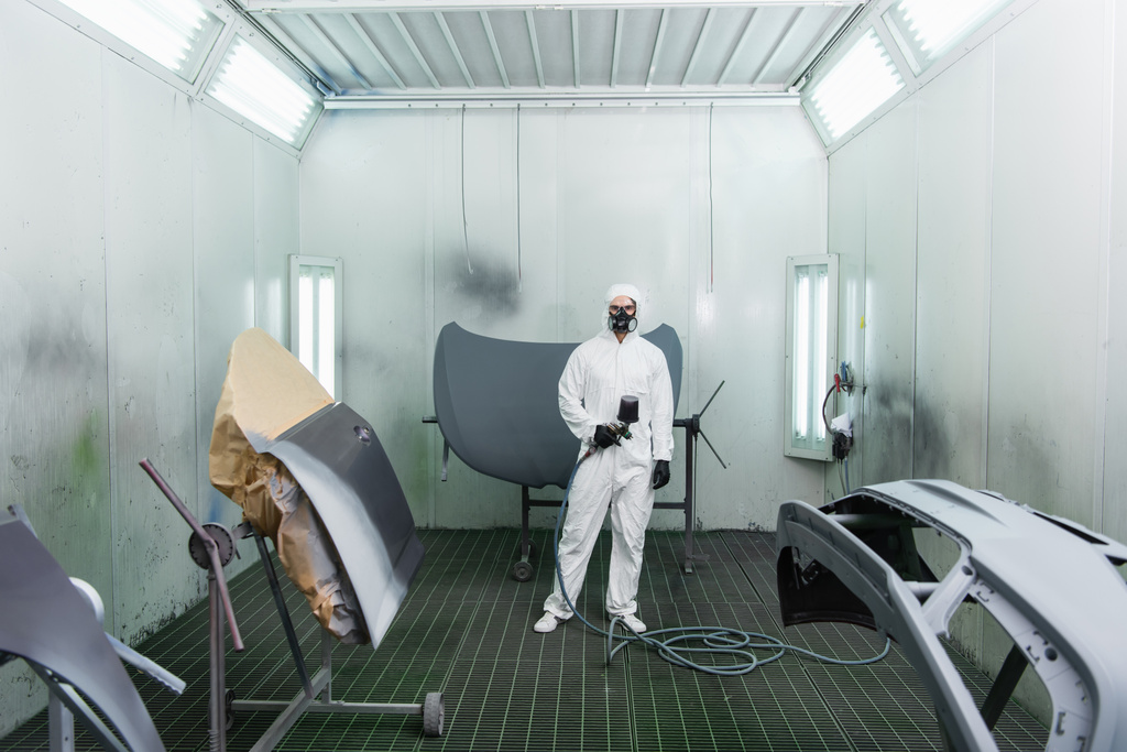 Munkás lélegeztetőgépben és vegyvédelmi ruhában aerográfot tart autóalkatrészek közelében a garázsban  - Fotó, kép