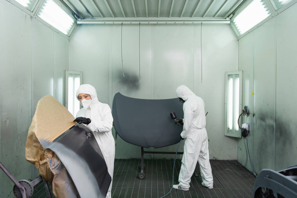 Jeune femme de travail en combinaison Hazmat et masque de protection debout près de la pièce de voiture tout en collègue utilisant aérographe dans le garage  - Photo, image