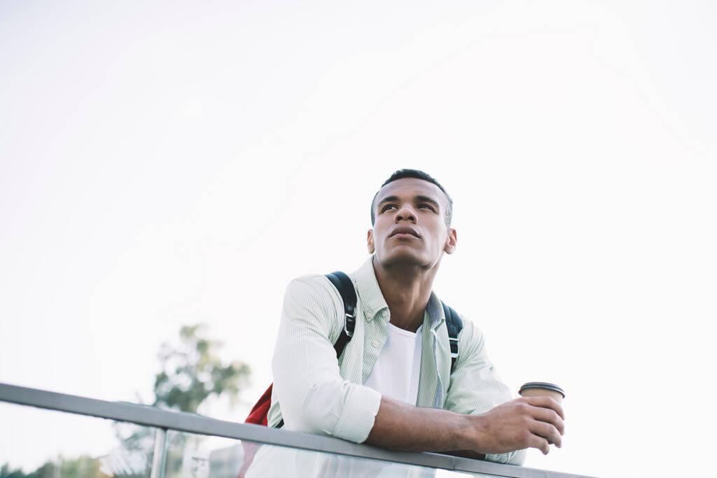 From below of thoughtful young black guy in casual clothes with backpack leaning on glass fence on city street with cup of drink in sunny summer day under cloudless sky - Foto, Imagem