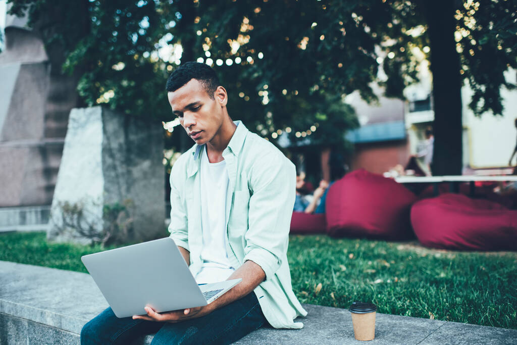 Young concentrated African American male in casual clothes sitting on concrete border in park and browsing laptop in summer evening - Foto, Bild