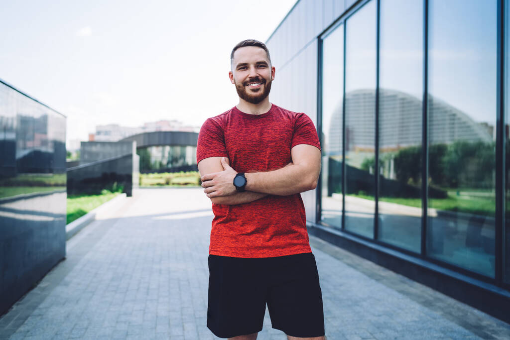 Positive male athlete in red tee shirt with wristwatch looking at camera and resting after workout while spending time in city - Foto, imagen