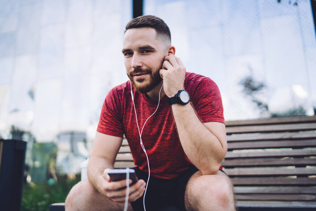 Молодий бородатий чоловік носить спортивний одяг за допомогою мобільного телефону, відпочиваючи на дерев'яній лавці на міській вулиці та насолоджуючись музикою
 - Фото, зображення