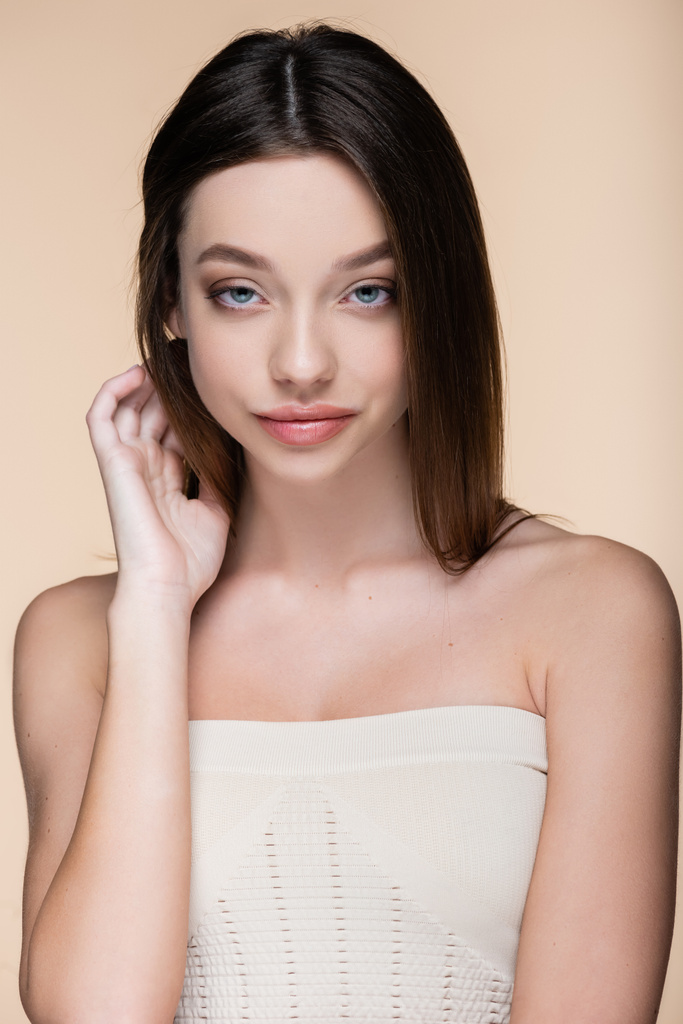 mujer joven con hombros desnudos ajustando el pelo aislado en beige - Foto, Imagen