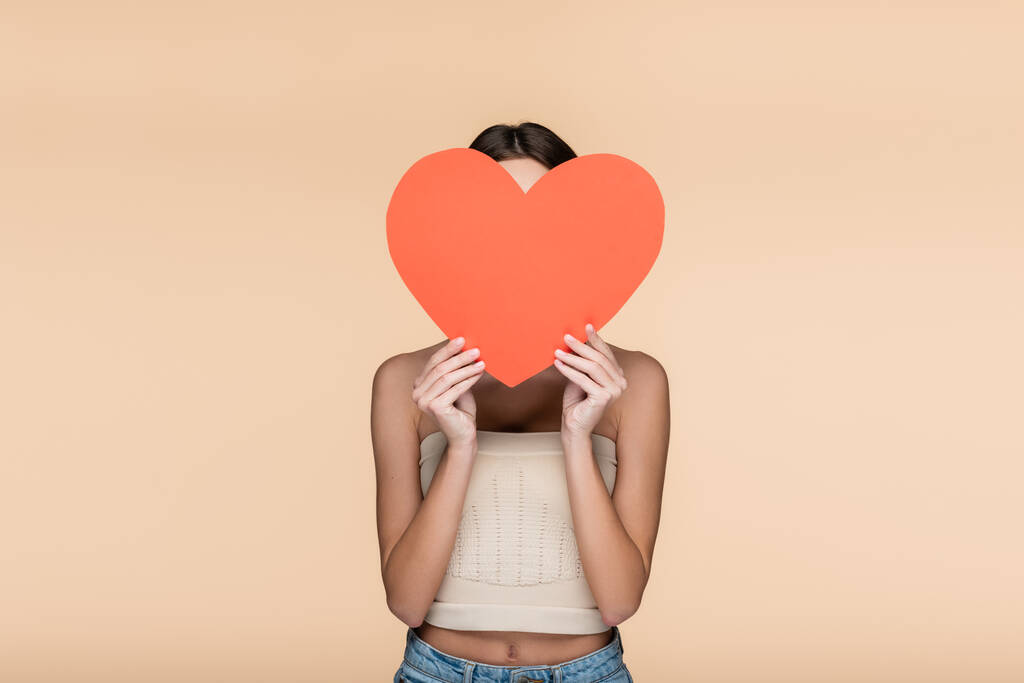 young woman in crop top with bare shoulders covering face with red paper heart isolated on beige  - Photo, Image