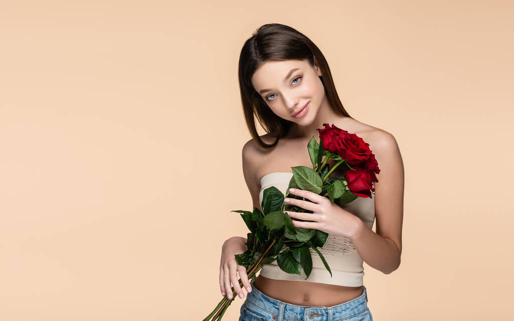 zufriedene junge Frau im Crop Top mit roten Rosen auf Beige  - Foto, Bild