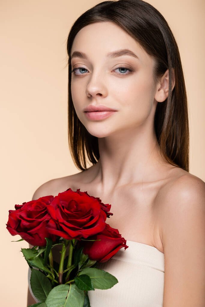 brunette young woman with bare shoulders holding red roses isolated on beige  - Fotó, kép