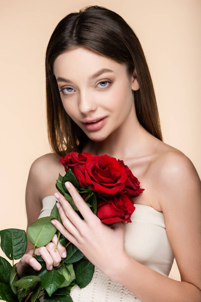 young woman with bare shoulders holding bouquet of red roses isolated on beige  - Фото, изображение