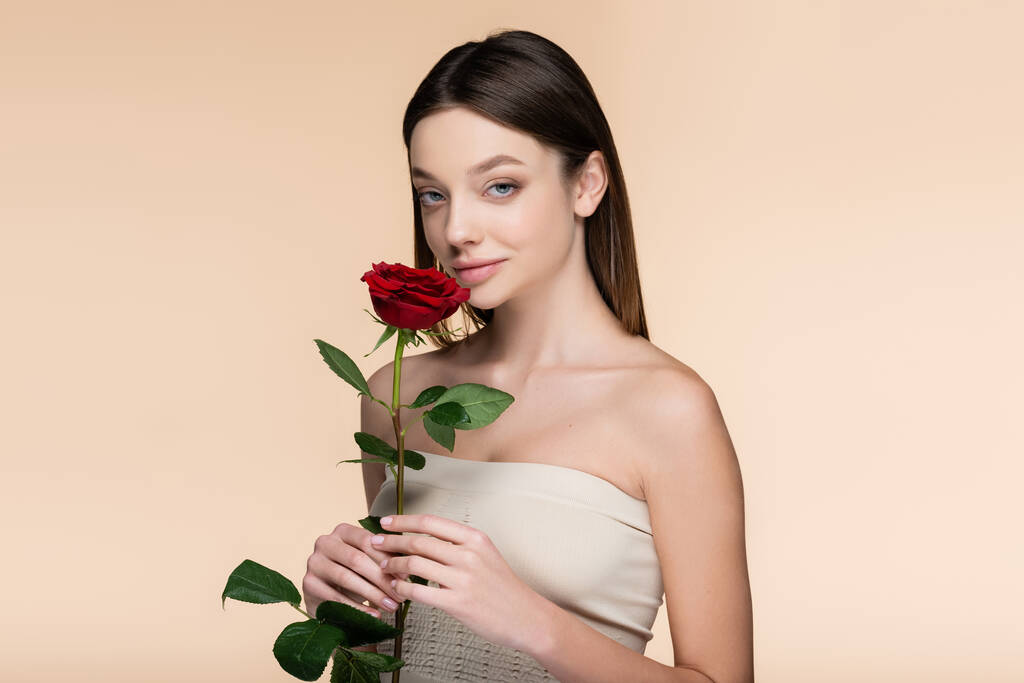 νεαρή γυναίκα με γυμνούς ώμους κρατώντας κόκκινο τριαντάφυλλο απομονωμένο σε μπεζ  - Φωτογραφία, εικόνα