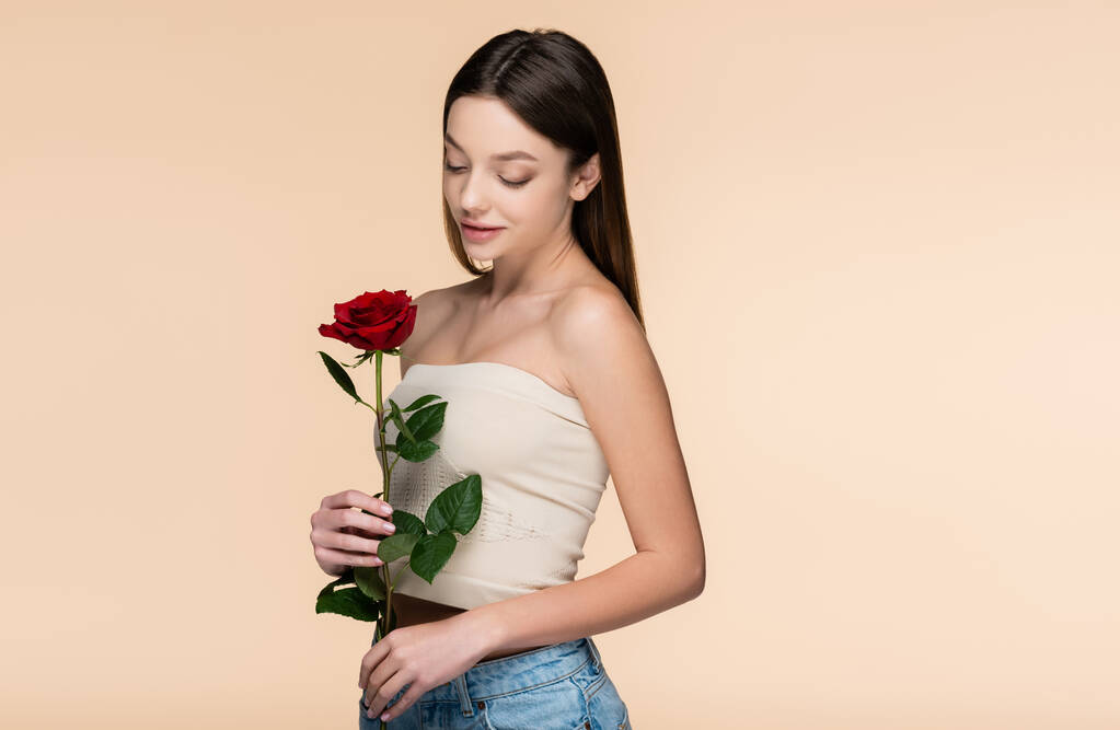 brunette jonge vrouw met blote schouders kijken naar rode roos geïsoleerd op beige  - Foto, afbeelding