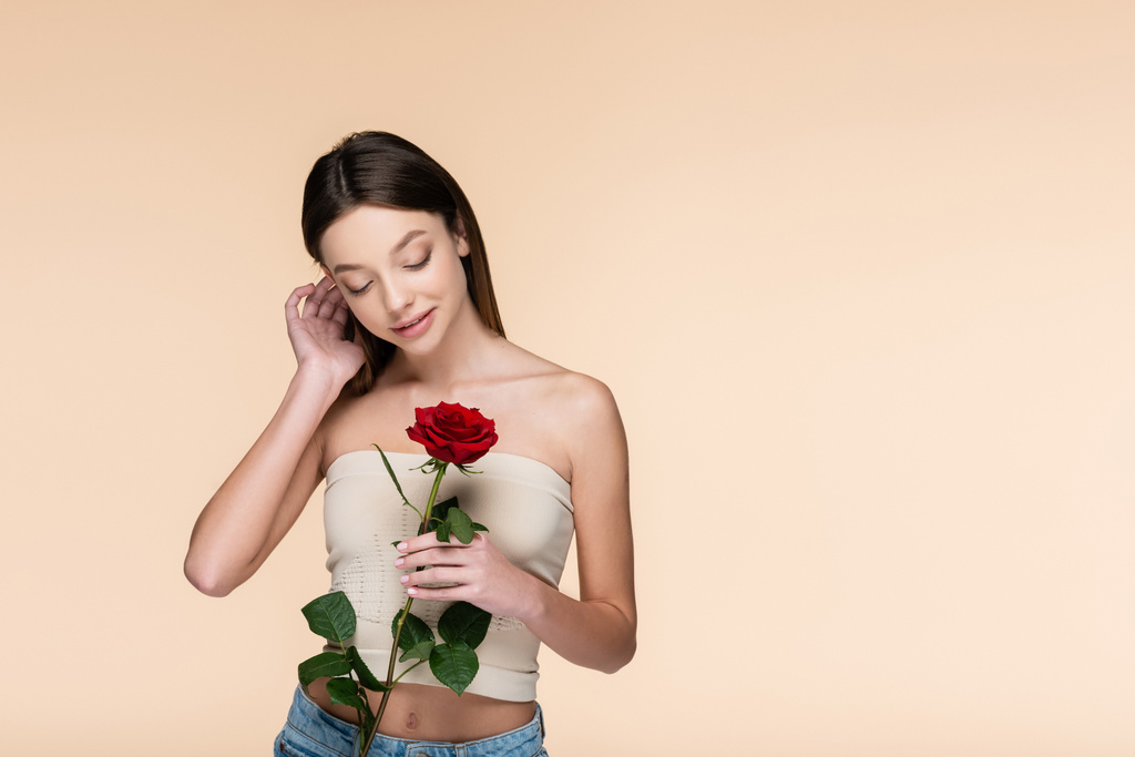 sensuele jonge vrouw met blote schouders kijken naar rode roos geïsoleerd op beige  - Foto, afbeelding
