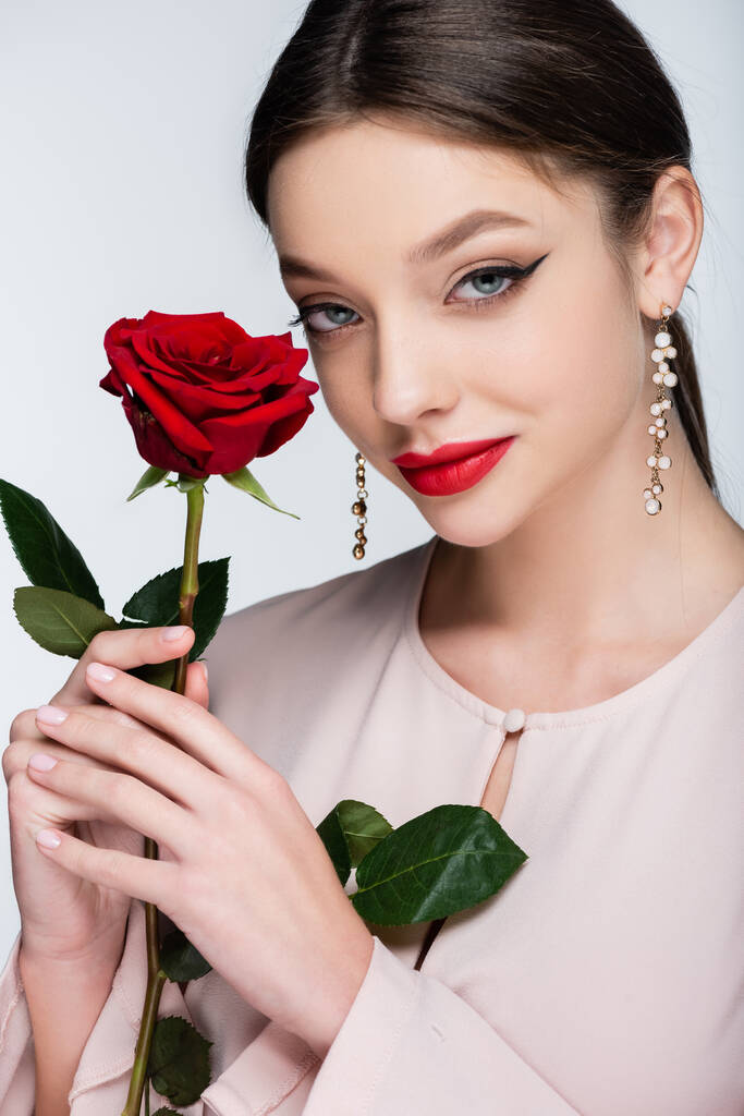 красивая молодая женщина в серьгах и блузке держа красную розу изолированы на серый - Фото, изображение