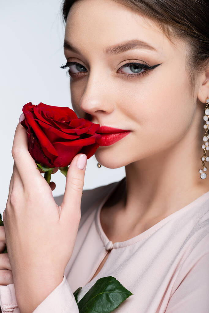 μελαχρινή γυναίκα με κόκκινα χείλη μυρίζοντας τριαντάφυλλο απομονωμένο σε γκρι - Φωτογραφία, εικόνα