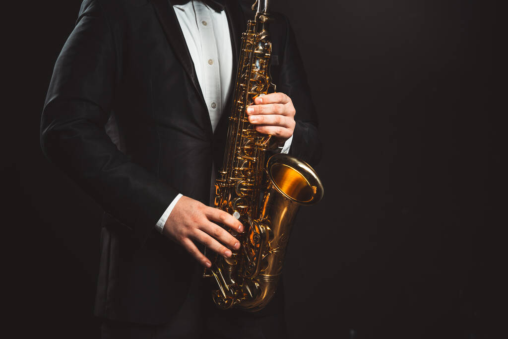 El saxofonista toca jazz. Músico masculino con un traje negro formal sostiene un saxofón tenor sobre un fondo oscuro. Primer plano del saxofón. Copiar espacio - Foto, imagen
