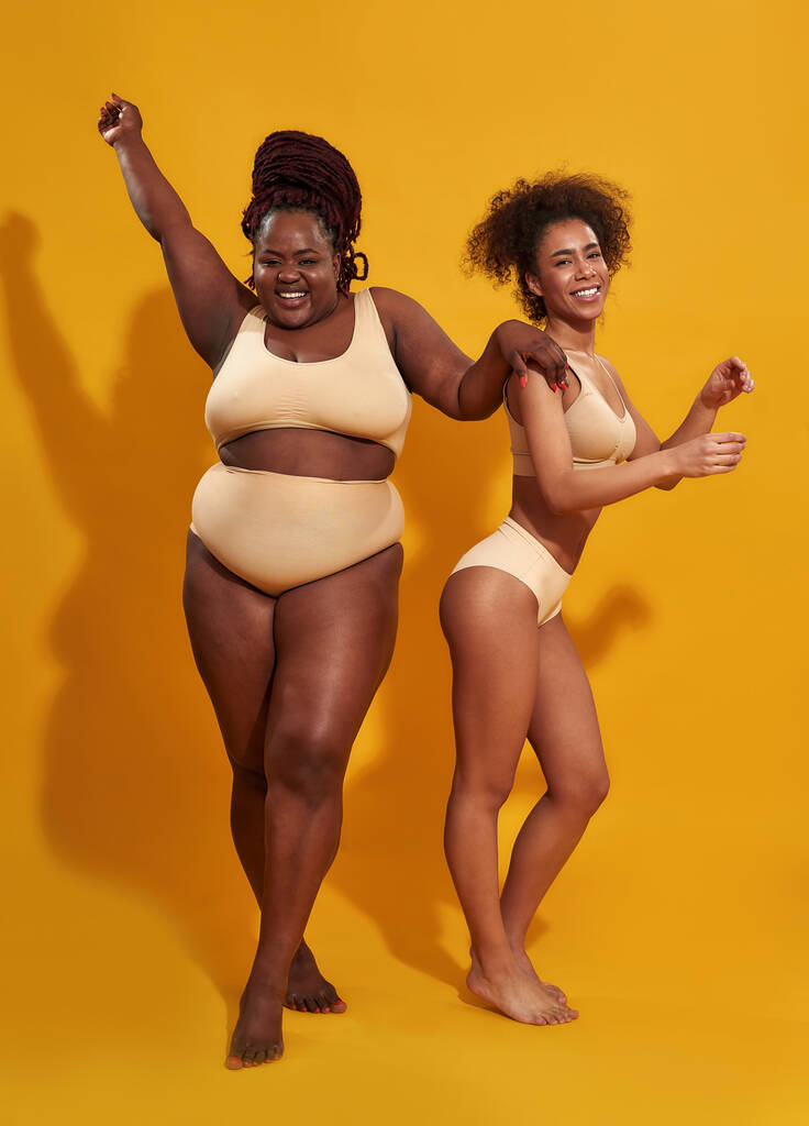 Dwie szczęśliwe, młode afro-amerykańskie kobiety w bieliźnie o różnym kształcie ciała uśmiechnięte do kamery, stojące razem na żółtym tle. - Zdjęcie, obraz