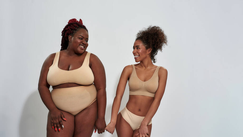 Twee vrolijke slanke en mollige Afrikaans-Amerikaanse vrouwen in beige ondergoed glimlachend naar elkaar, samen staan geïsoleerd over grijze achtergrond - Foto, afbeelding