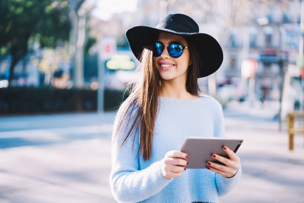 Chica hipster vestida de moda con gafas de sol elegantes y sombrero con almohadilla táctil moderna de pie en la calle de la ciudad y sonriente, bloguera femenina feliz con tableta digital o ebook disfrutando de un día soleado en vacaciones - Foto, imagen