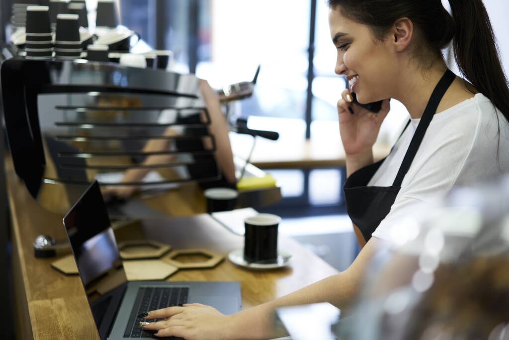 Positive female owner of small business cafe using devices for organizing job making call to banking service operator, smiling waitress talking on cellular with clients reserving table in coffee shop - Foto, imagen