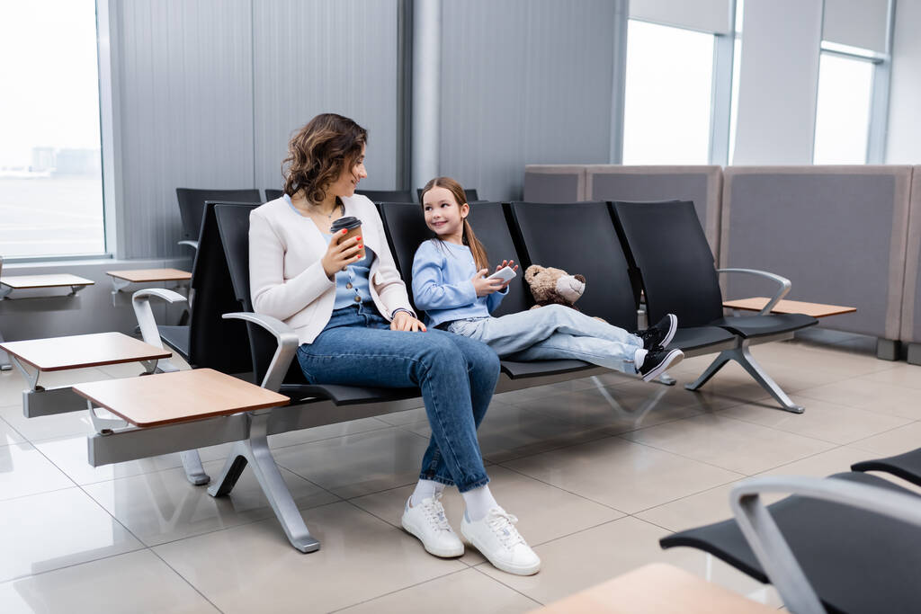 niño alegre sosteniendo teléfono inteligente y mirando a la madre con taza de papel en el salón del aeropuerto - Foto, Imagen