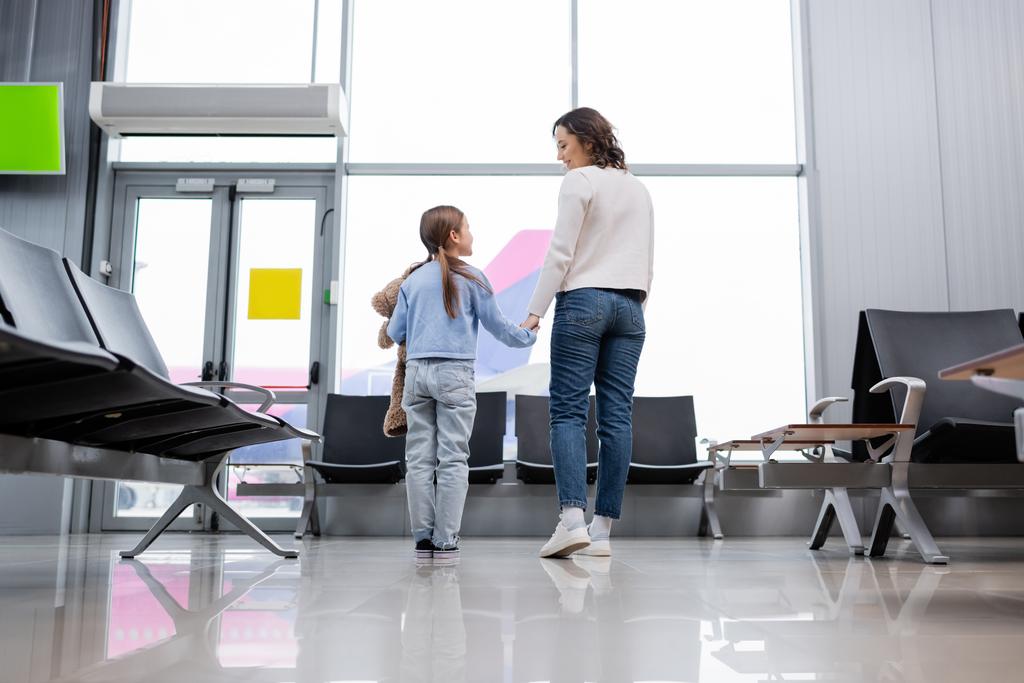 feliz madre e hija tomados de la mano mientras se miran en el salón del aeropuerto  - Foto, Imagen