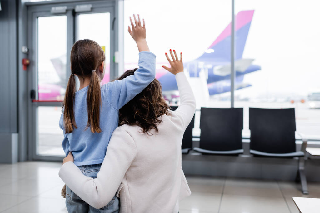 back view of mother and daughter waving hands while looking at plane in airport  - Photo, image