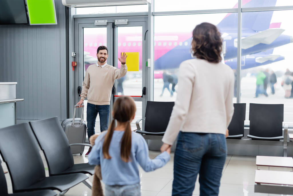 Χαρούμενος άνθρωπος με αποσκευές χαιρετώντας το χέρι, ενώ κοιτάζοντας τη γυναίκα και την κόρη στο αεροδρόμιο  - Φωτογραφία, εικόνα