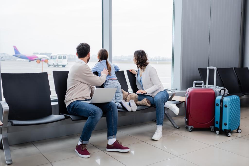 man and woman holding gadgets while sitting near daughter and looking at plane through window in airport lounge  - Foto, Imagem