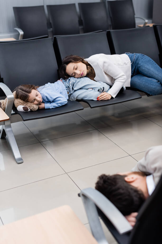 мати і дочка сплять на сидіннях аеропорту біля розмитого чоловіка в залі вильоту
  - Фото, зображення