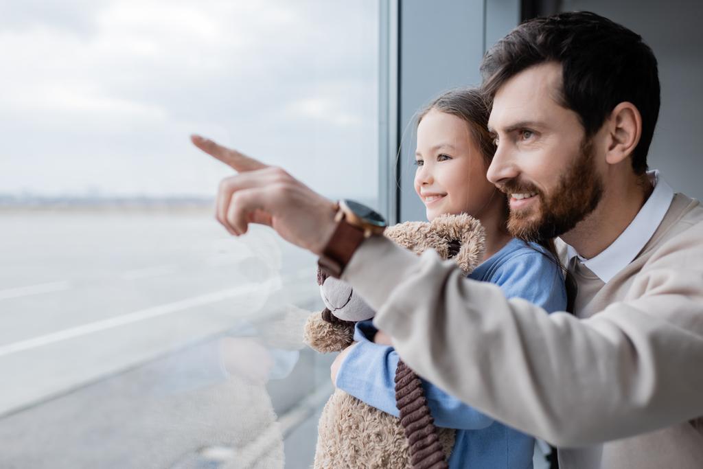 happy man pointing with finger at window near daughter with soft toy in airport - Foto, Imagen