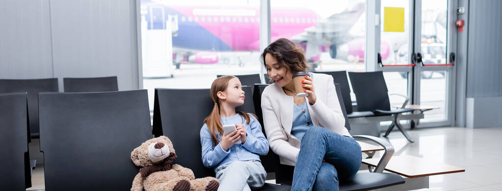 niño sosteniendo teléfono inteligente y mirando a la madre alegre con taza de papel en el salón del aeropuerto, pancarta - Foto, Imagen