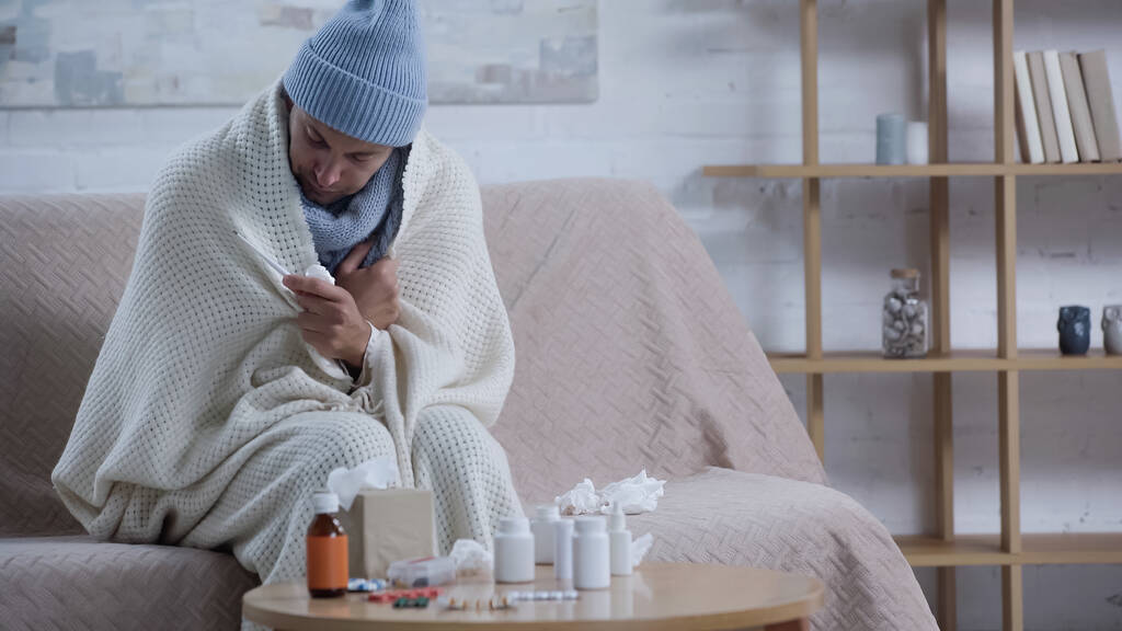 hombre enfermo sentado en el sofá en manta caliente y sombrero cerca de la mesa con servilletas de papel y medicamentos - Foto, imagen