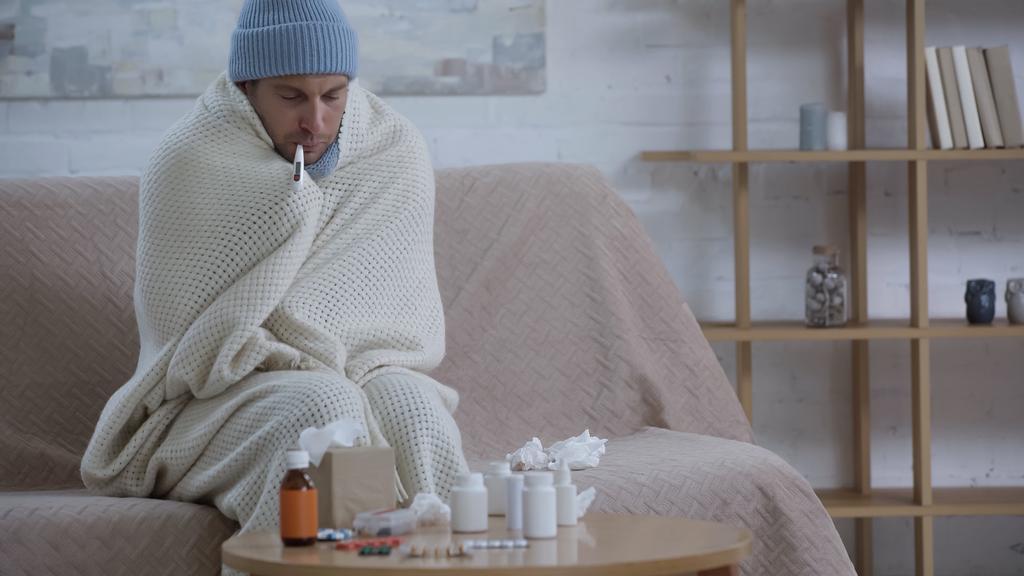 sick man in warm hat and beanie measuring temperature near table with medicaments - Foto, imagen