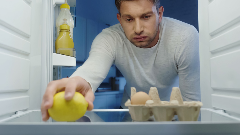displeased man puffing cheeks while taking lemon from refrigerator - Fotó, kép