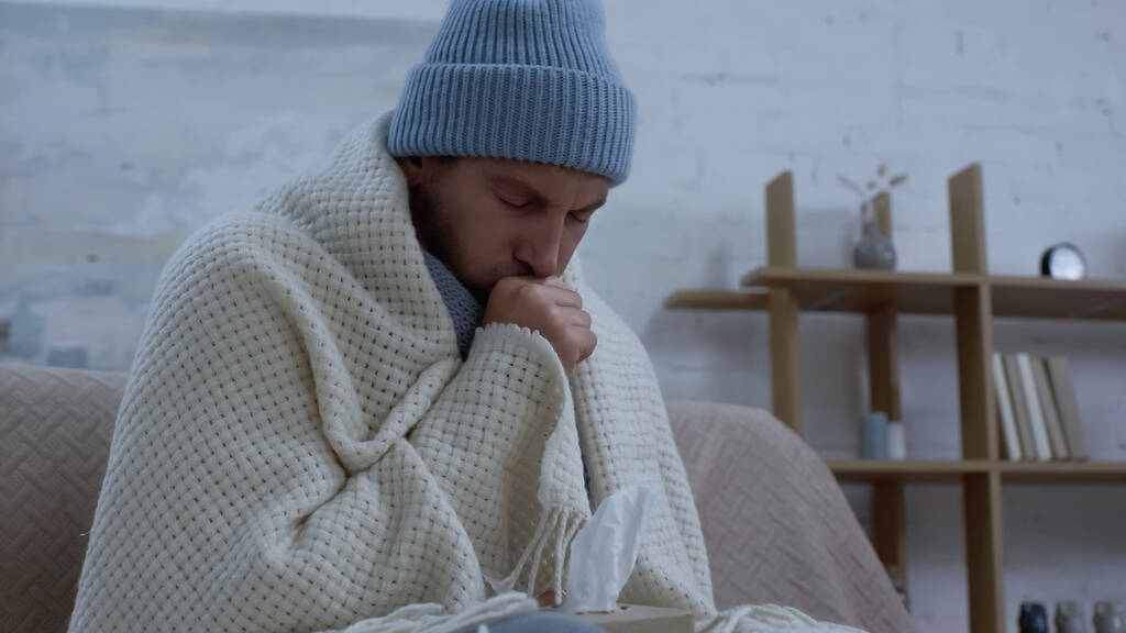 άρρωστος άνδρας με ζεστό καπέλο και κουβέρτα βήχει στον καναπέ στο σπίτι - Φωτογραφία, εικόνα