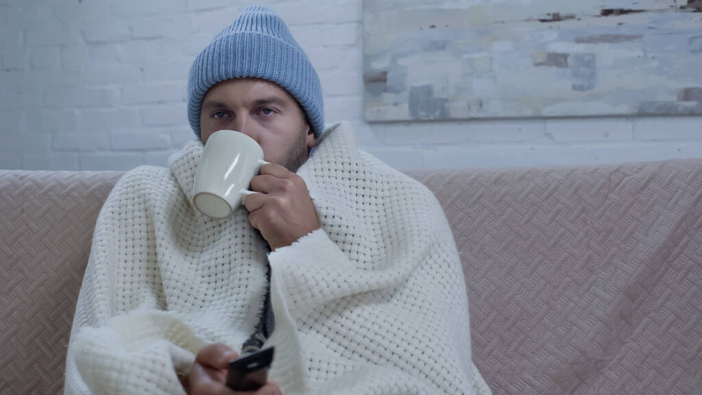 sick man drinking warm beverage and watching tv while sitting on sofa in beanie and blanket - Foto, afbeelding
