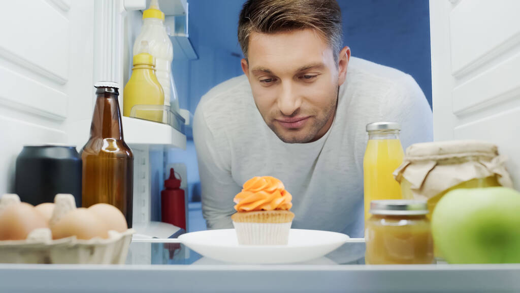 man looking at delicious cupcake near drinks, eggs and sauces in fridge - Фото, изображение