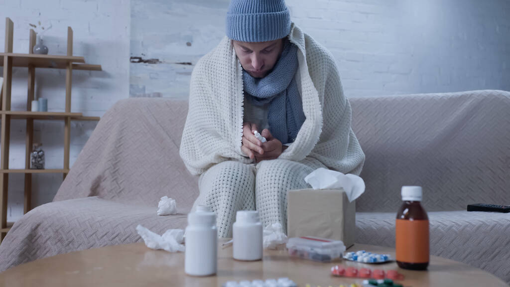 sick man in warm hat, scarf and blanket holding nasal spray near table with medication and paper napkins - Foto, Imagen